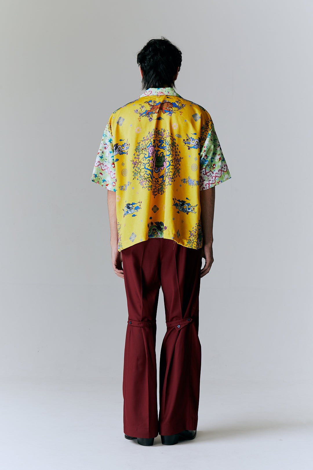 Person wearing a bright yellow floral shirt and burgundy pants on a plain background